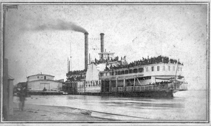 An original photograph shows the Sultana steamship in port, loaded with passengers. Smoke rises from the funnels. Passengers gleefully hold their hats in the air as a farewell gesture.   