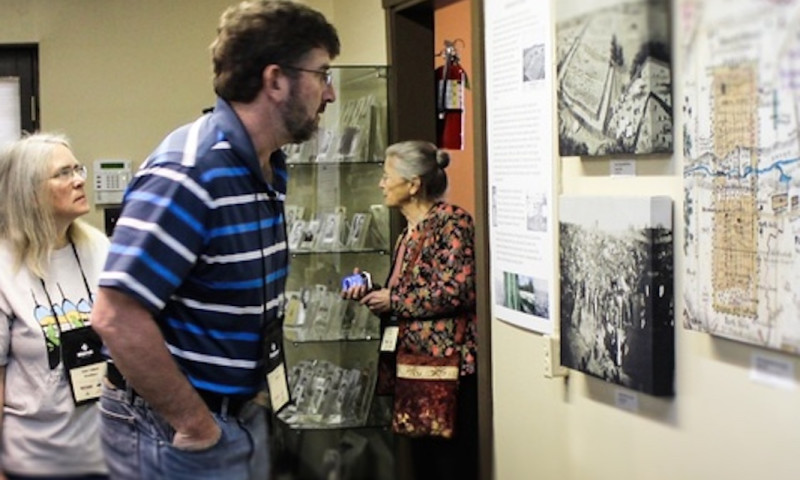 A group of people is looking at various artifacts in the Sultana Disaster Museum. Maps and images cover the walls. A glass display case contains standing photos.