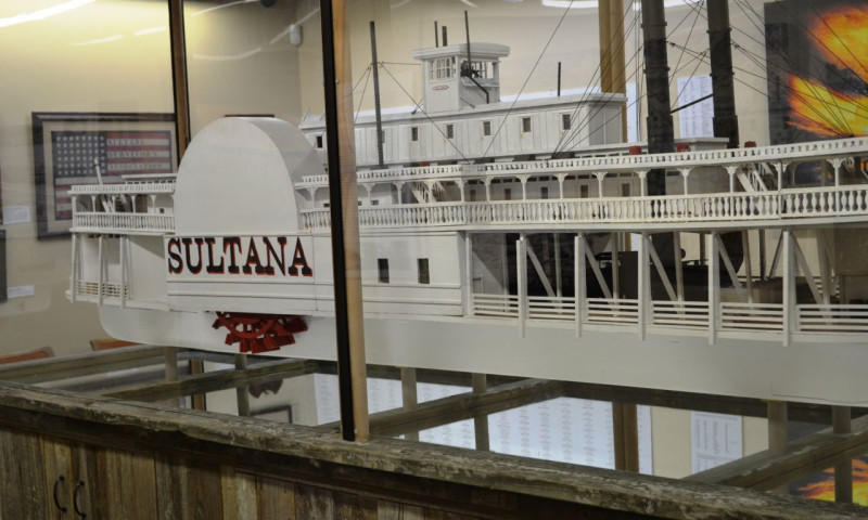 A room-sized replica of the steamship Sultana is presented behind a glass partition. A bright red paddlewheel is visible. Above it, red and black letters denote “Sultana.” The ship is white, with intricate railings, cables and other details visible.