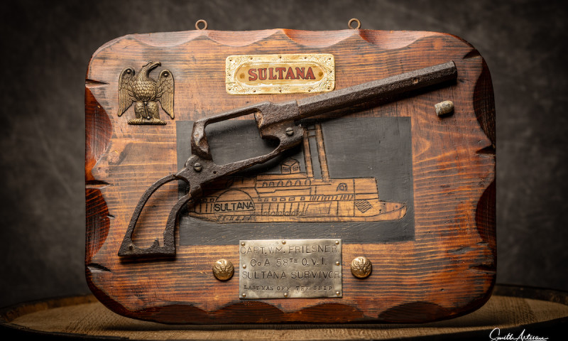 The rusted frame of a handgun is mounted on a wooden background bearing the name and likeness of the Sultana. A plaque describes the gun as once belonging to Captain William Friesner, noted as the “last man off the ship.”