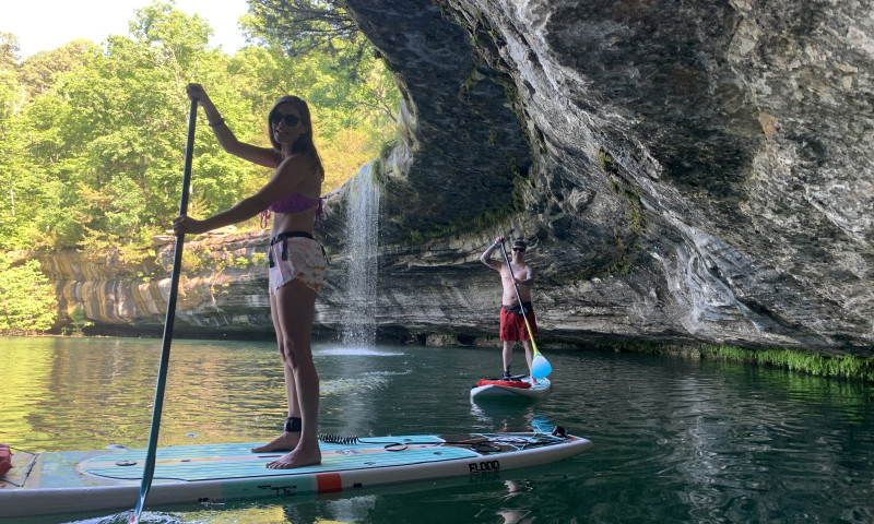 A lake setting with a limestone bluff and waterfall. A girl is standing on a paddleboard next to the bluff. A boy can be seen standing on a board behind her. 
