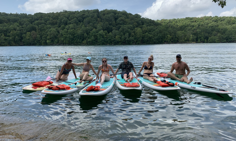 A lake setting. A group of six people in a range of ages sit on paddleboards that face the camera. 