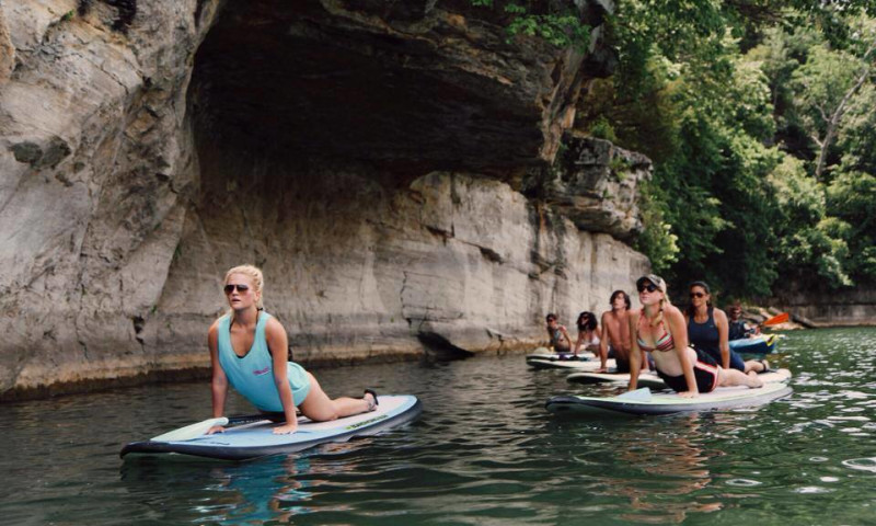A lake setting with a bluff to the side. A group of 5 to 7 paddleboarders are doing a cobra pose modeled after one instructor who is at the front of the group. 
