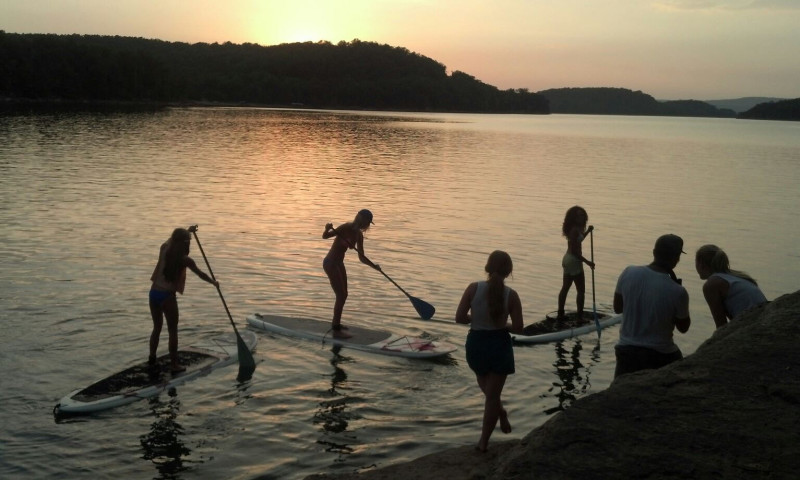 A lake setting at sunset. A trio of people are on paddleboards. Three other people are on the bank in the foreground. 