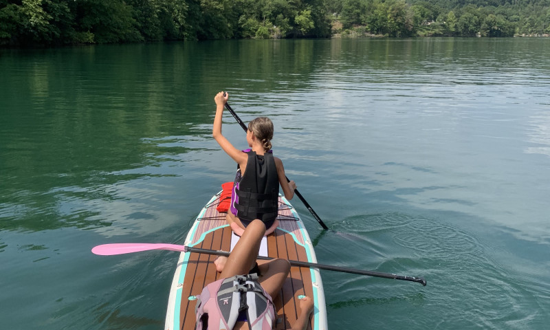 A lake setting with a bank of trees in front. A girl sitting down on a paddleboard with her back facing camera. A second girl can be seen laying down on the paddleboard in back. 
