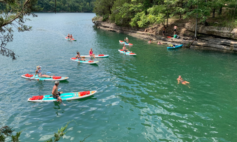 A lake setting with a rock and tree bank on the right side of frame. A group of paddleboarders are in the water. Kayaks are pulled up to the banks. 