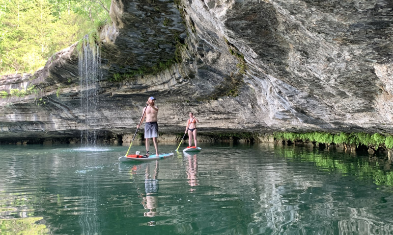 Waterfalls ArkansasA lake setting that is enveloped by a limestone bluff. A man and a woman are standing on their paddleboards, floating through the water. 