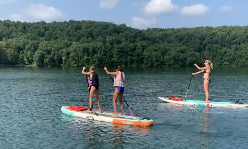 A lake setting. Two girls wearing life vests are on a paddleboard together on the left. A woman in a bikini is on a board on the right. Both are facing away from the camera. 