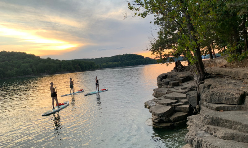 A lake setting. The sun is setting over a line of trees. A bank of rocks and trees is at the right of the frame. Three paddleboarders are standing up facing the sunset. 