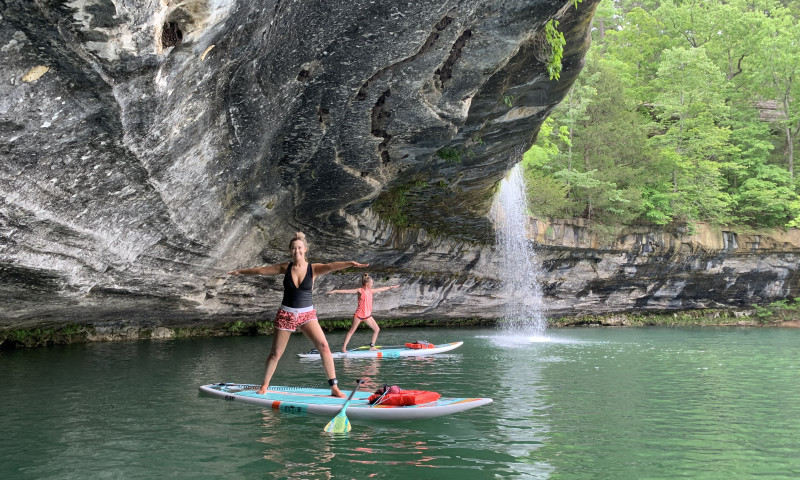 A lake setting with a limestone bluff in the background. Two women are standing on paddleboards, starting to do a warrior yoga pose. 