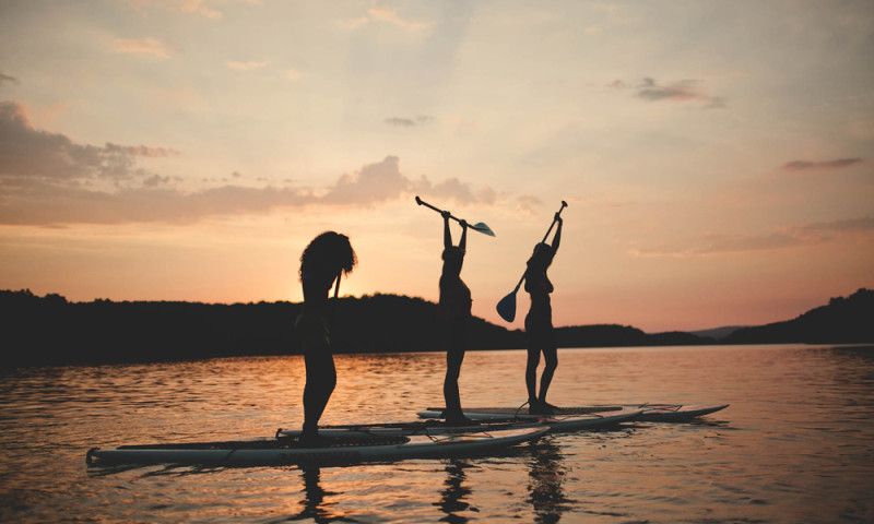 A lake setting at sunset. Three women stand on paddleboards with oars raised in hand.