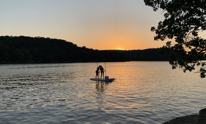 A lake setting at sunset. Trees are in the foreground and background. Two people can be seen standing up and kissing on paddleboards. 