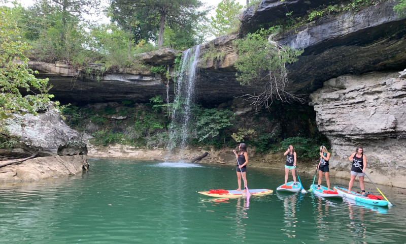 A lake setting with bluffs and a waterfall surrounding the water. Four women in matching shirts stand up on paddleboards. 