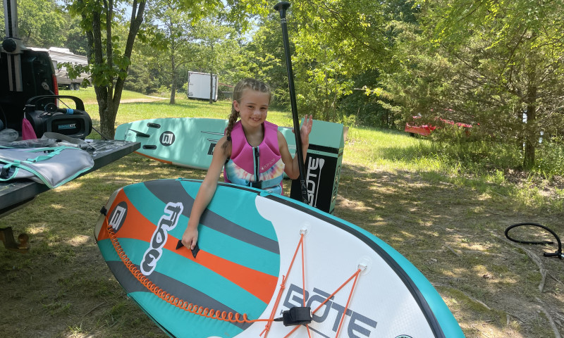 A wooded, grassy setting. A young girl holds a paddle and a paddleboard while she stands next to a tailgate. 