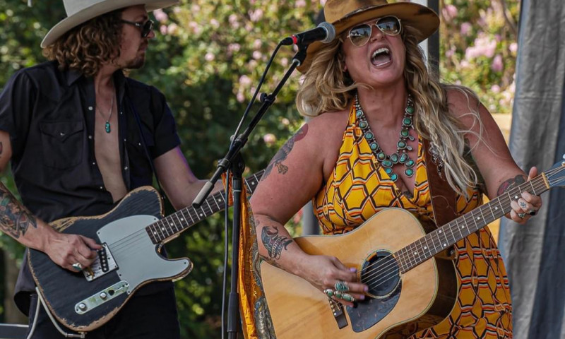 Woman wearing yellow dress and hat playing acoustic guitar and singing into a microphone onstage next to a man in a hat playing electric guitar.
