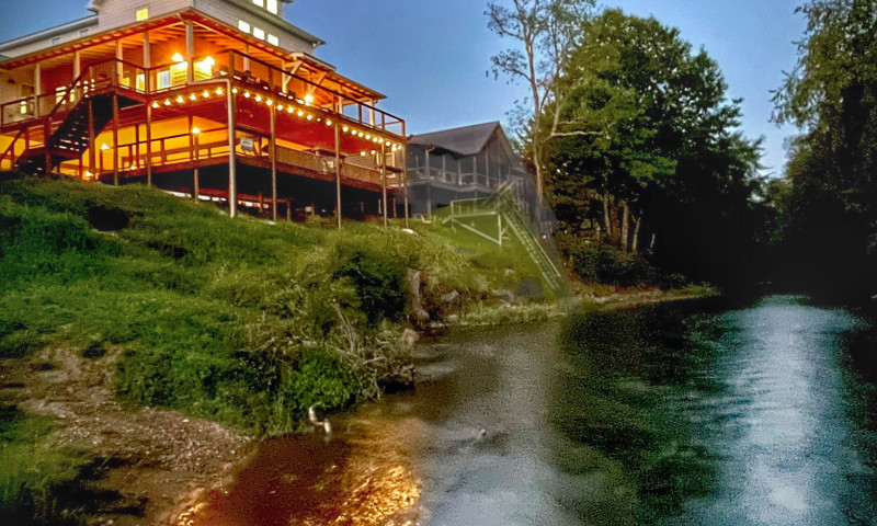 Evening view of a beautiful riverside cabin brightly illuminated by porch lights.