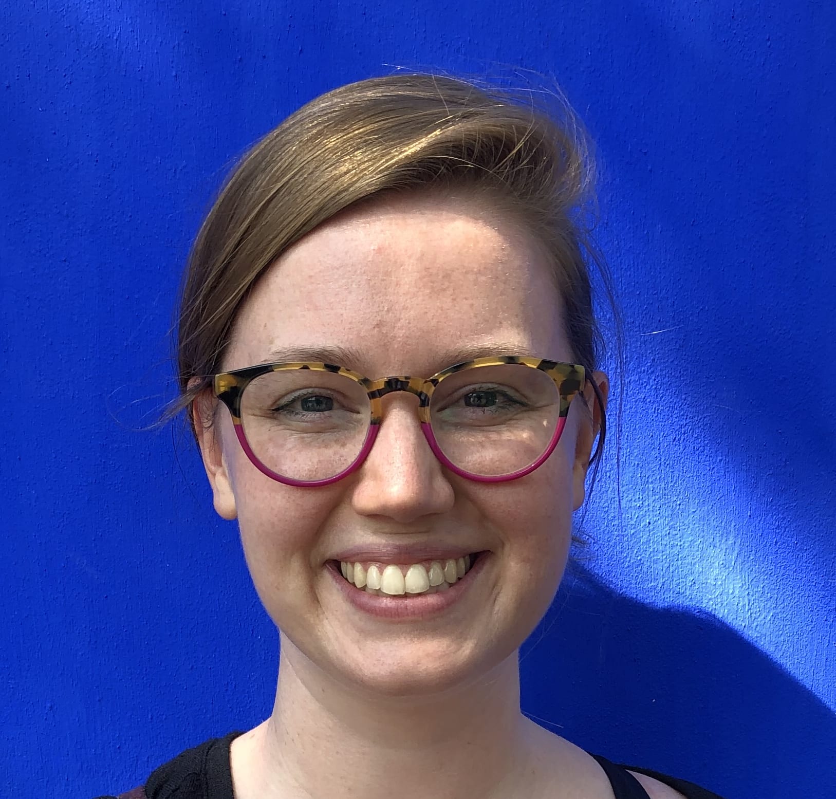 A photo of me, Taylor: a smiling woman wearing glasses against a bright blue wall.
