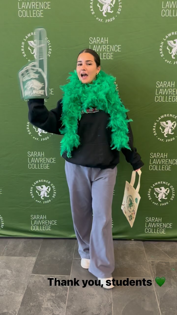 Person with a green feather boa lifting a trophy, standing in front of a college banner.