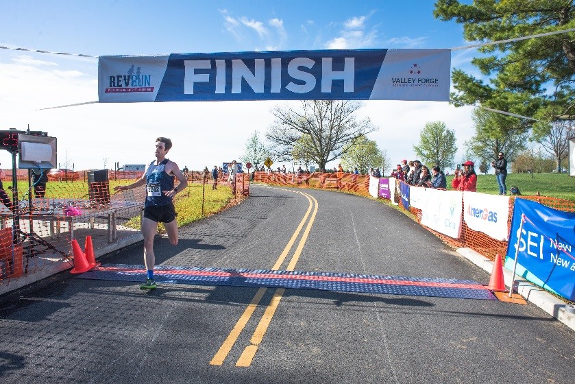 First-place winner Trevor van Ackeren of Bethlehem blasts across the finish line to complete the 2017 Valley Forge Revolutionary 5-Mile Run®. He completed the course in 25:28.