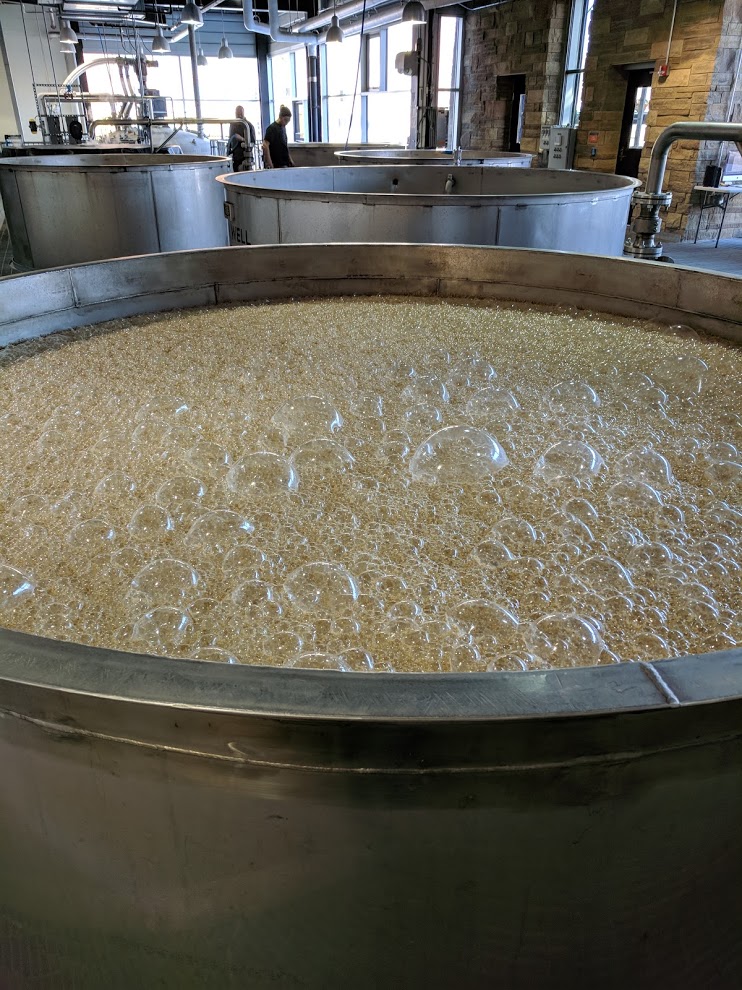 A huge mash tub with brown bubbling whiskey mash in the foreground and a couple of silver metal tubs in the background at New Riff Distilling