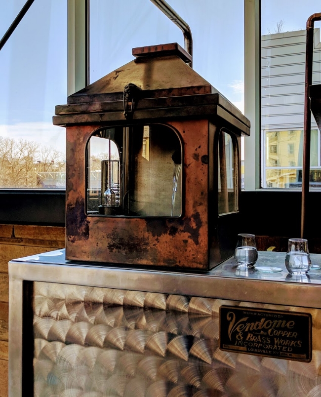 Copper spirit safe with new make bourbon visible through the glass window at New Riff Distillery