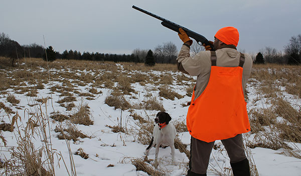 Hunter wearing reflective gear aiming shotgun in the air with his trusty dog in the snow