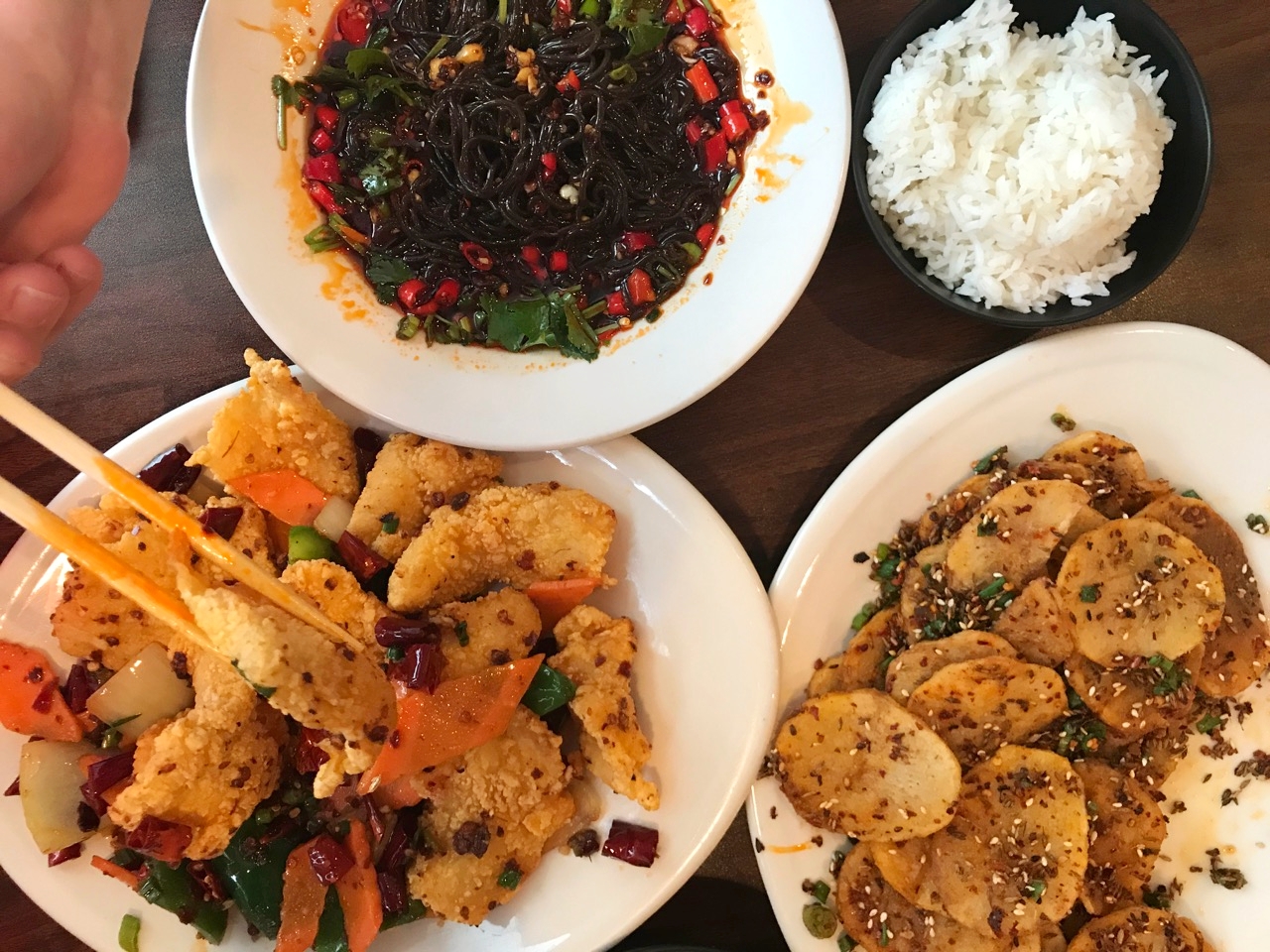 Meal from NE Chinese including multiple plates of fried meat & veggies, black noodles, rice, and fried eggplant