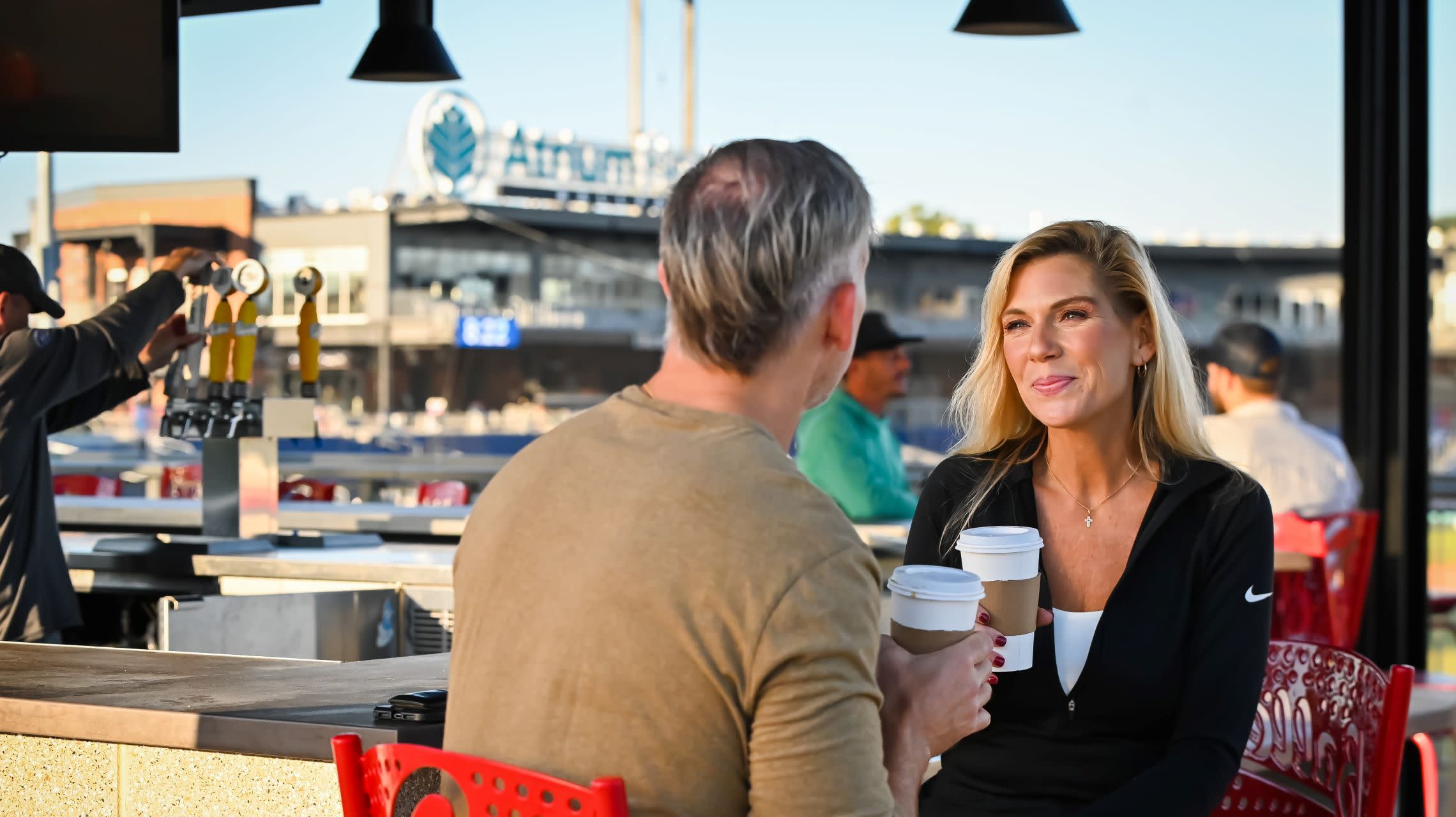 man and woman sit and drink coffee at bar located near ballpark outfield