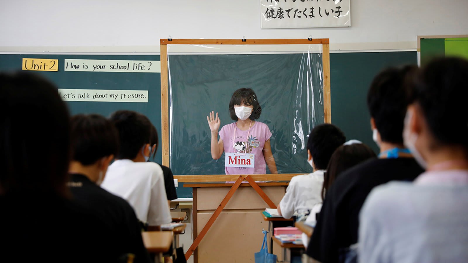 A kid stands in front of a school classroom, behind a COVID protection screen and a mask.