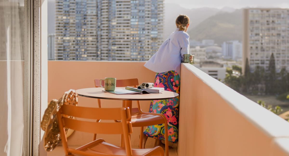 A person gazing out over downtown Waikīkī from a private lanai. There is a table and two chairs on the lanai set with coffee mugs and a magazine.