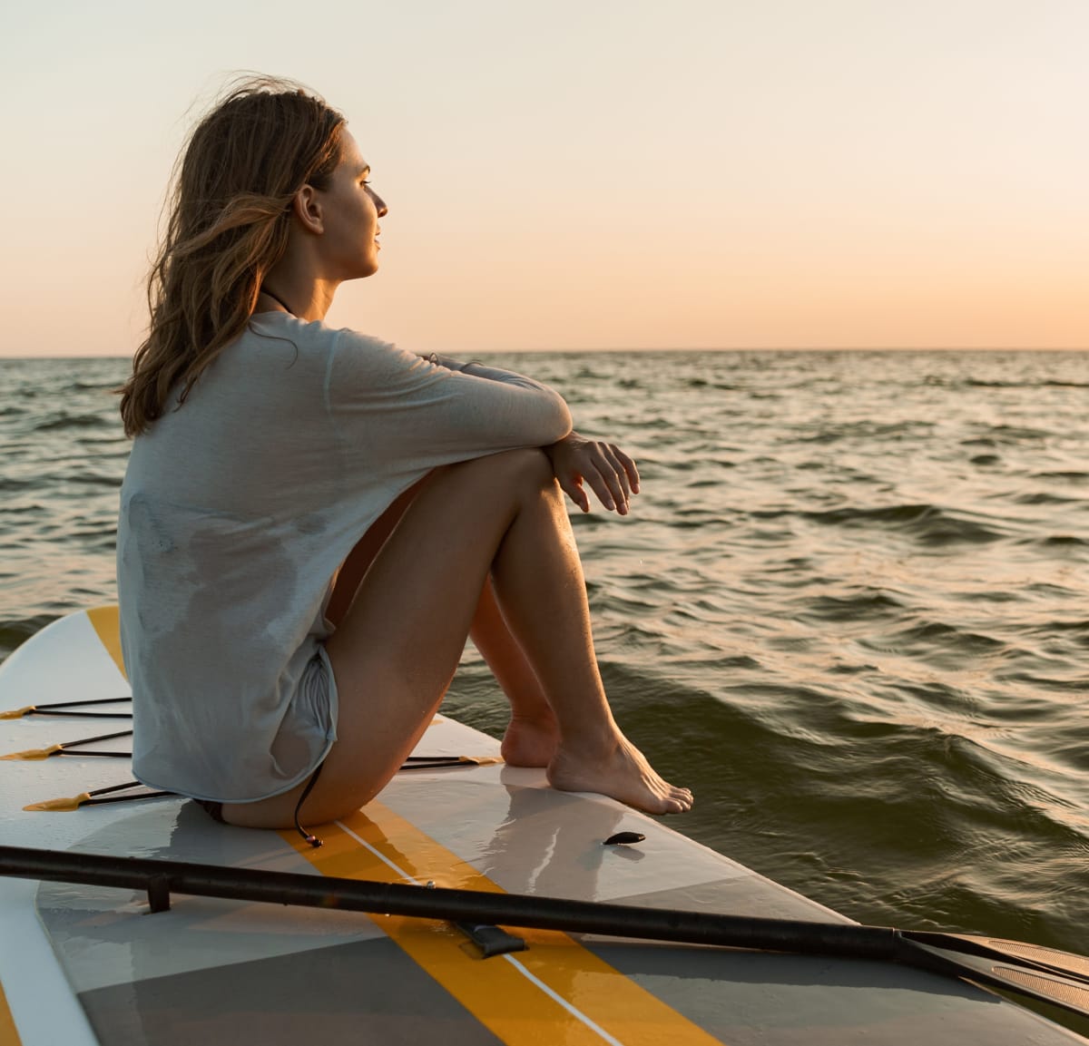 Girl sitting on paddleboard on ocean looking at sunset
