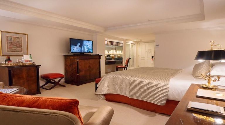 This photo showcases our 400 square foot room, featuring one King size bed against the side wall with two night stands on each side. Across from the bed there is an armoire with dresser drawers and a 43 inch flat screen TV. Next to the armoire you will find a fully stocked mini bar, including wine, bottled beverages and assorted snacks available for purchase. Located near the window there is a comfortable armchair with an additional side table next to it. The room also features a full size work desk, an Italian marble bathroom with a 55 gallon soaking tub and a vanity.