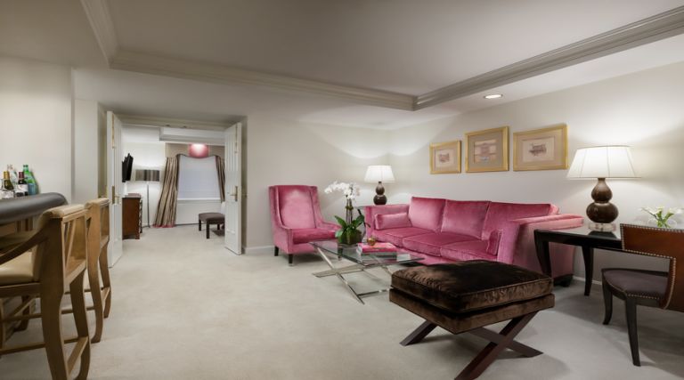 This photo showcases our 600 square foot suite, featuring either a Queen size bed with a sitting bench at the foot of the bed. French doors separate the bedroom and living room. The living room includes a seating area with one armchair, a full size sofa, and ottoman. In the center of the lounge area is a glass coffee table with matching end table with lamp. Adjacent to the seating area is a full size bar with marble counter top and three bar stools. . Wall decor includes Italian inspired artwork. The bathroom is made with Italian marble and features a 55 gallon soaking tub.