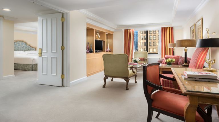 This photo showcases our 600 square foot one bedroom suite, with a King size bed against the wall and a seating bench at the foot of the bed. Across from the bed is a custom made wooden wall unit, featuring six drawers, glass shelving with books, and a 43 inch flat screen TV. This elegant wall unit also features two sets of french doors leading into the living room. Next to the bed is a lounge chair with a standing lamp. Opposite the bed is the full size Italian marble bathroom with pocket doors. The living room features a seating area with a sofa, armchair, coffee and side table. The armoire in the living room is equipped with an additional 43 inch flat screen TV.