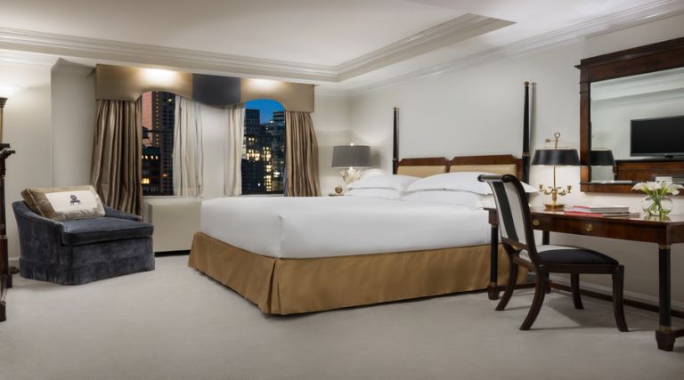 This photo showcases our 450 square foot room, featuring one King size bed against the side wall with two night stands on each side. Across from the bed there is an armoire with dresser drawers and a 43 inch flat screen TV. Next to the armoire you will find a fully stocked mini bar, including wine, bottled beverages and assorted snacks available for purchase. Located near the window there is a comfortable armchair with an additional side table next to it. The room also features a full size work desk, an Italian marble bathroom with a 55 gallon soaking tub and a vanity.