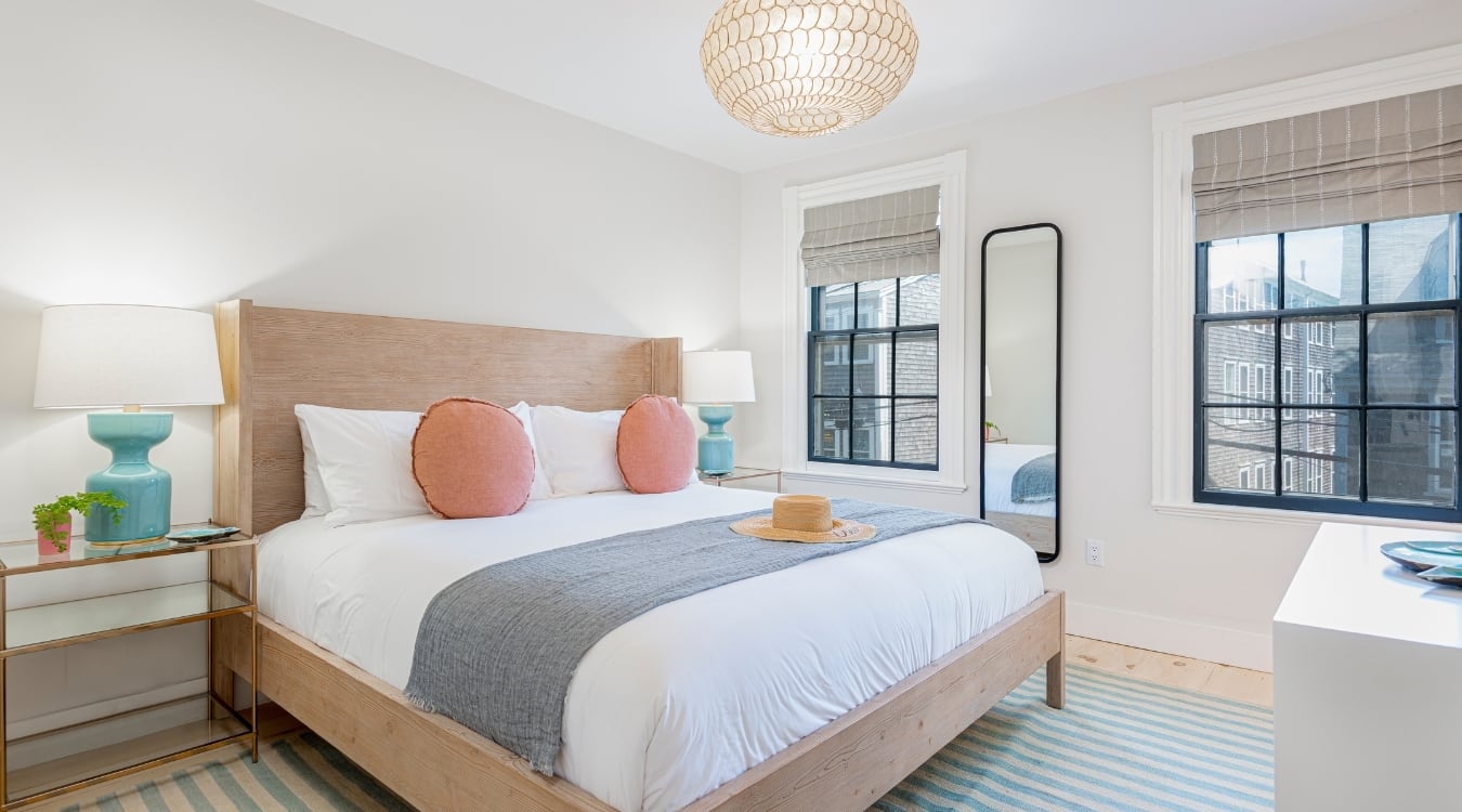 To the left we have a bed with pillows and a hat placed on it. Lamps are placed beside the bed. A mirror is fixed to the wall between two windows.