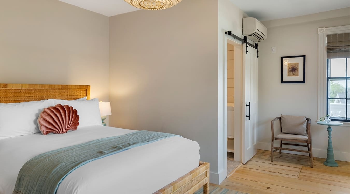 To the left we have a bed with table lamp beside it. To the right, we have a chair and a table stand with a vase placed on it. AC is fixed above the wardrobe door.