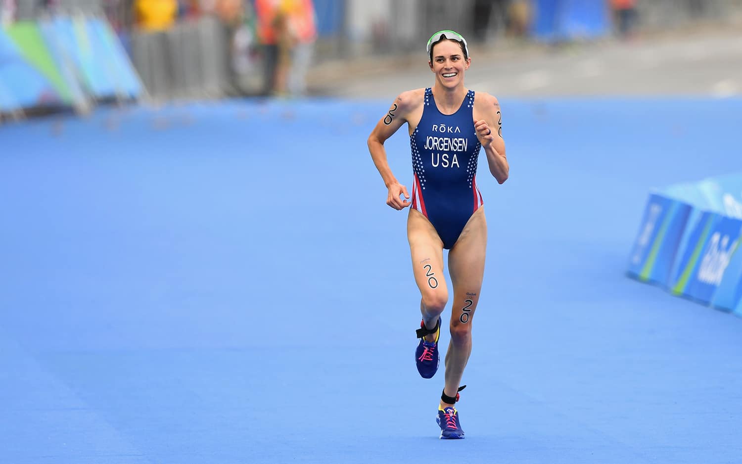 Triathlete Gwen Jorgensen runs to the finish line of the 2016 Olympics. She is wearing a red, white and blue tri kit and is smiling.