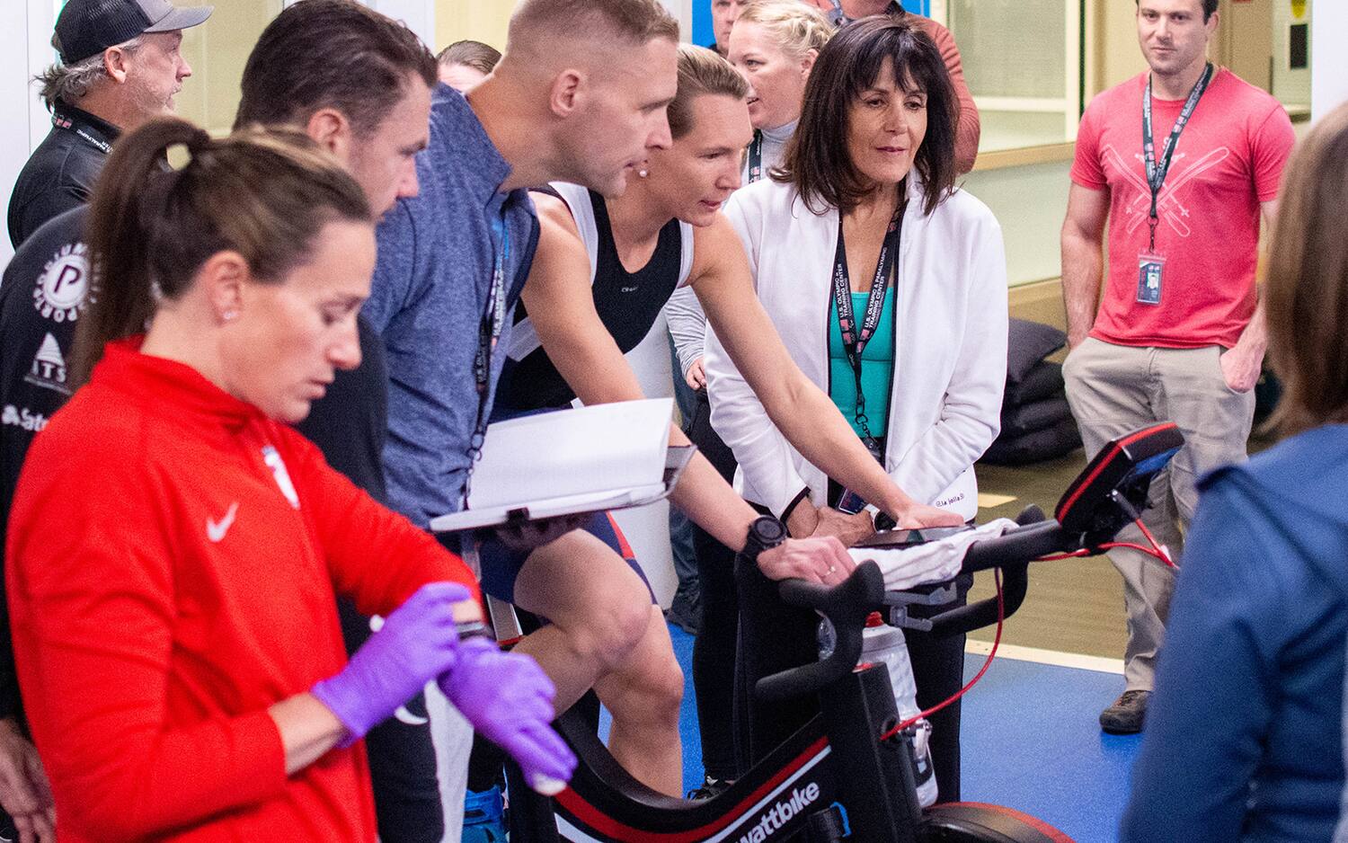 Coaches watch a power meter as an athlete rides an indoor bike trainer. In the foreground, an instructor checks her watch.