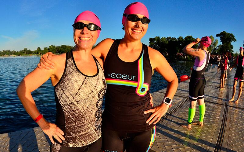 two women wearing trisuits stand on the dock before a triathlon race, with their arms around each other