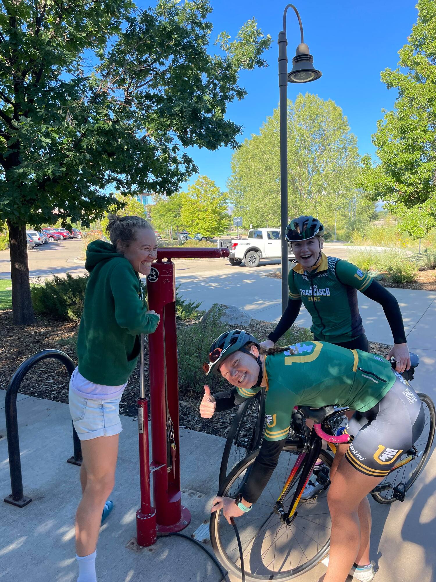 two female triathletes work on filling air in the tires of a bike and third triathlete is smiling and laughing