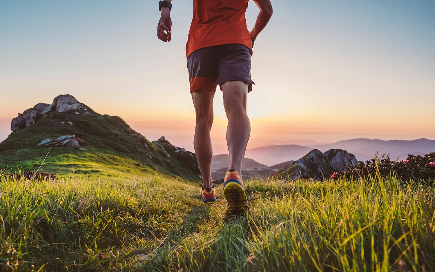 An athlete runs on grass towards a group of rocks. The sun is either rising or setting in the background.