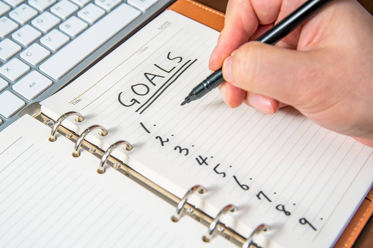 A hand holding a pen is shown writing in a notebook. The page's header is