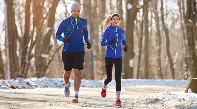 Two athletes run during  on a road with snow lining the edges. Both are dressed for cold weather with gloves, headgear, and long sleeves.