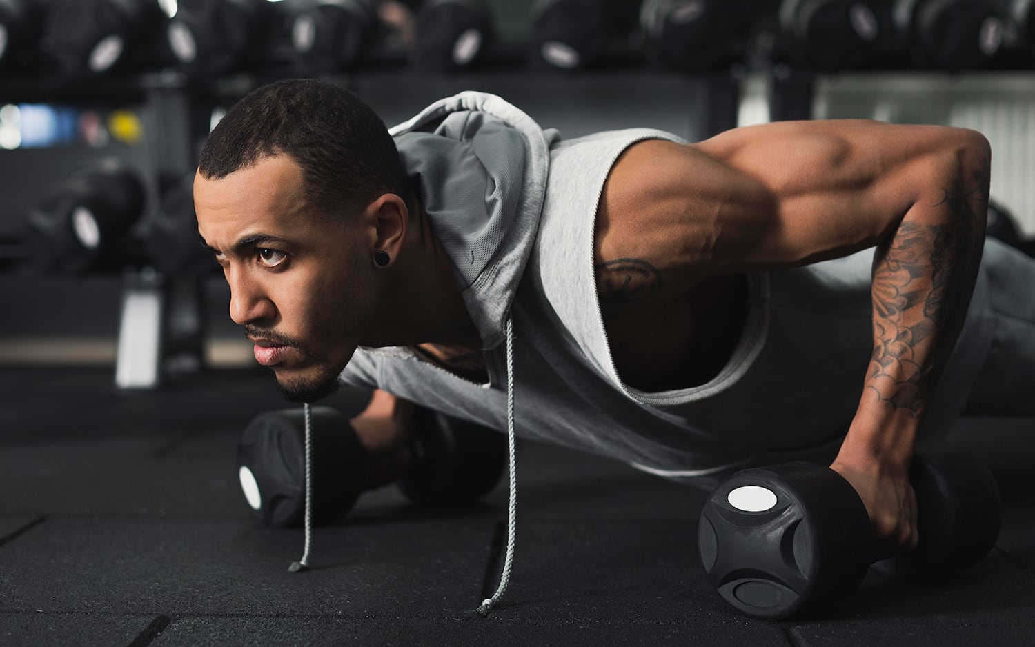 An athlete lies in a pushup position while holding a dumbbell in both hands. He has a determined look on his face.
