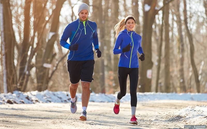 Two runners bundle up while running outside with snow and ice surrounding the road.