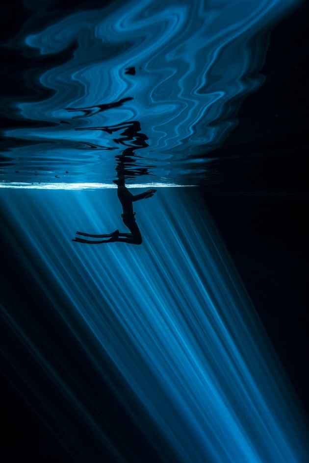 © Wendy Timmermans, Netherlands. WINNER - PRIMARY COLOURS (SINGLE IMAGE CATEGORY). Cenote Nah Yah, Mexico. As freedivers, we explore the abyss on one breath only. Inside the Cenote the water was completely still and silent, but his tiny movements on the surface created an irregular pattern of reflection, beautifully contrasting with the straightness of the rays underwater.