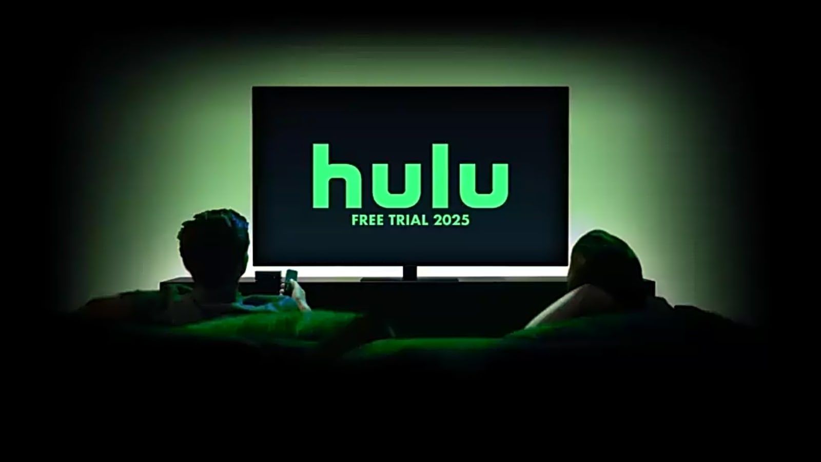 The Hulu logo on a flat screen TV in the dark with two people on the couch watching with the verbiage "Free Trial 2025" beneath the logo on the flat screen