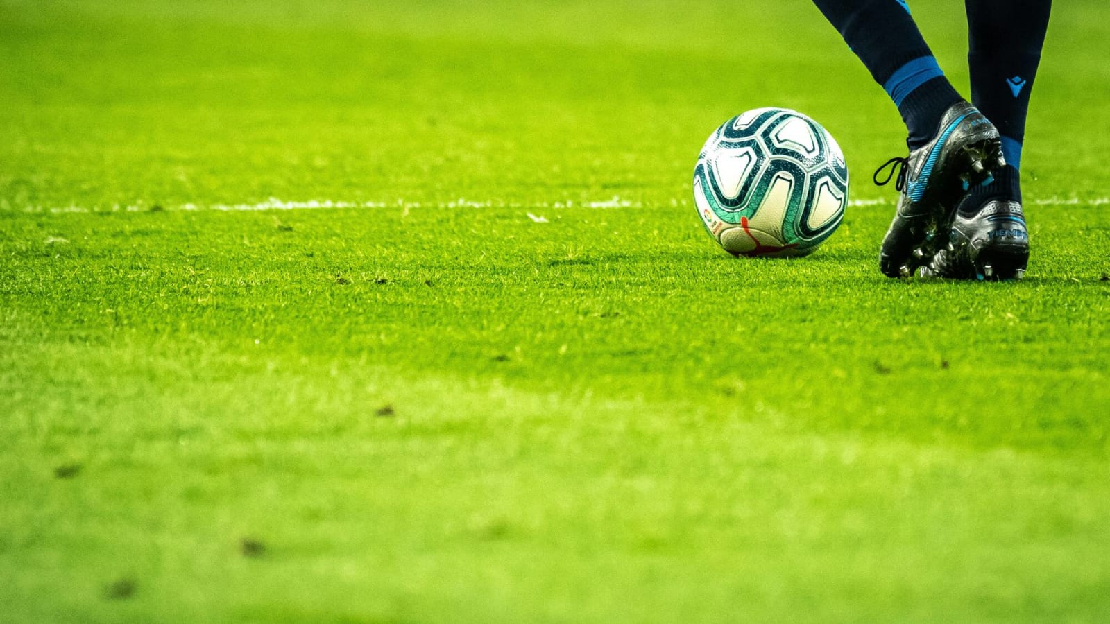 The lower half of a man's body wearing blue cleats about to kick a teal soccer ball on a grassy field