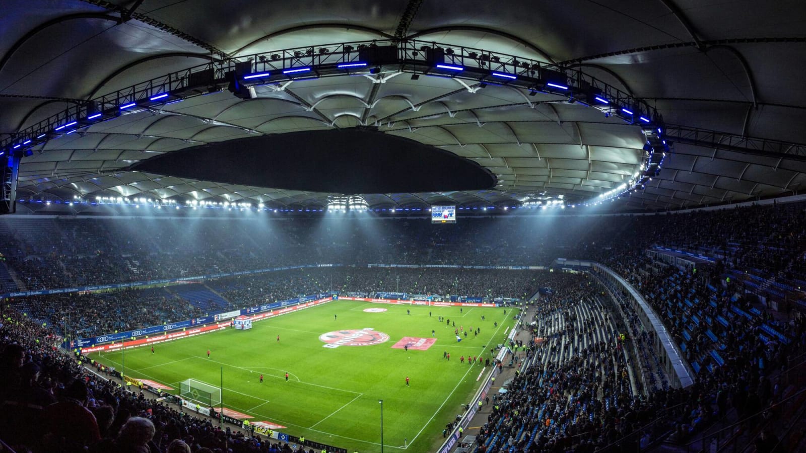 A covered soccer stadium with players under lights just taking the pitch